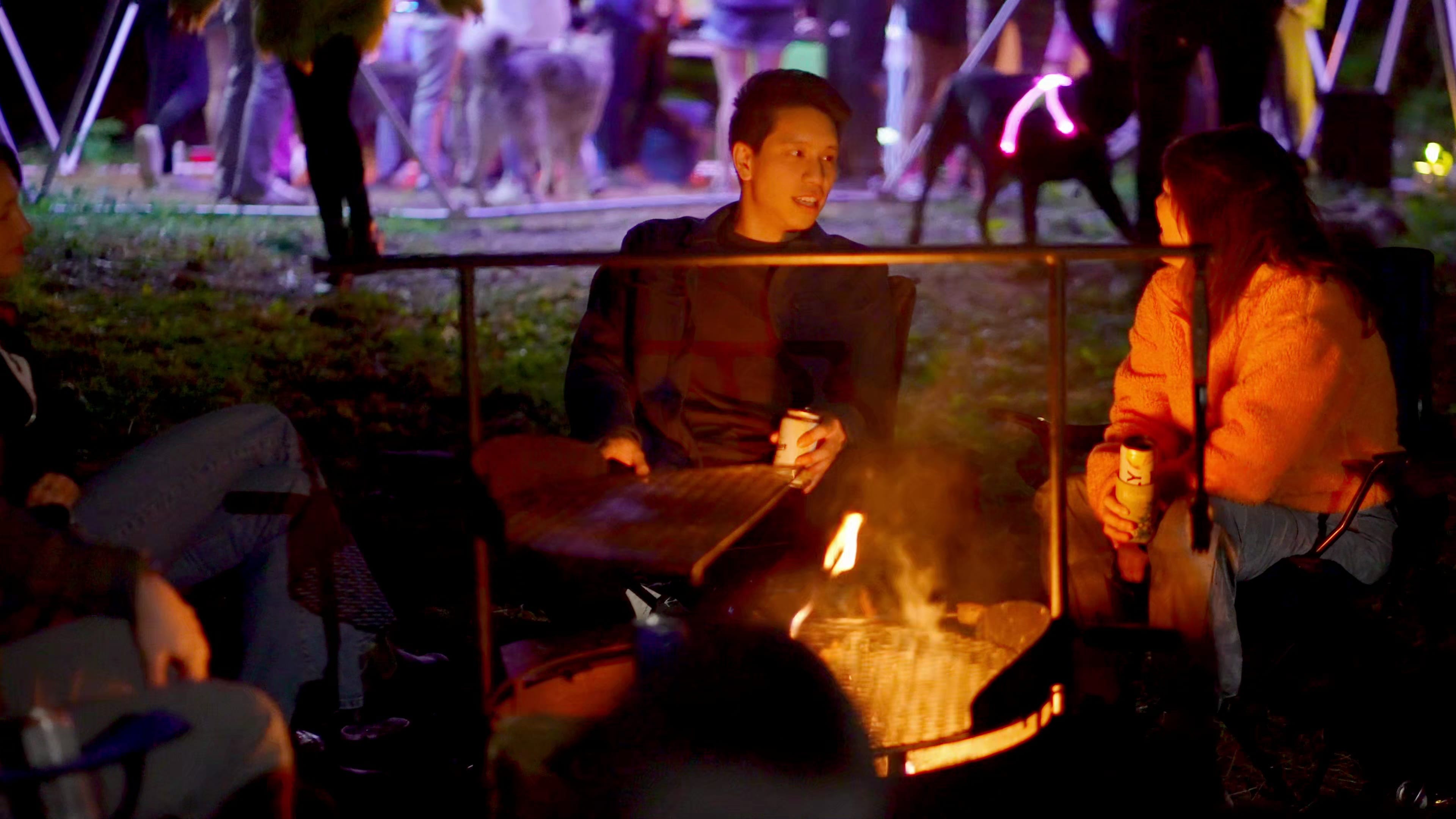 Friends gathered around a firepit at night with neon lights in the background